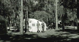 a historical photo in black and white shows a man and woman walking in front of a camping tent in the trees. the man is holding a baby.